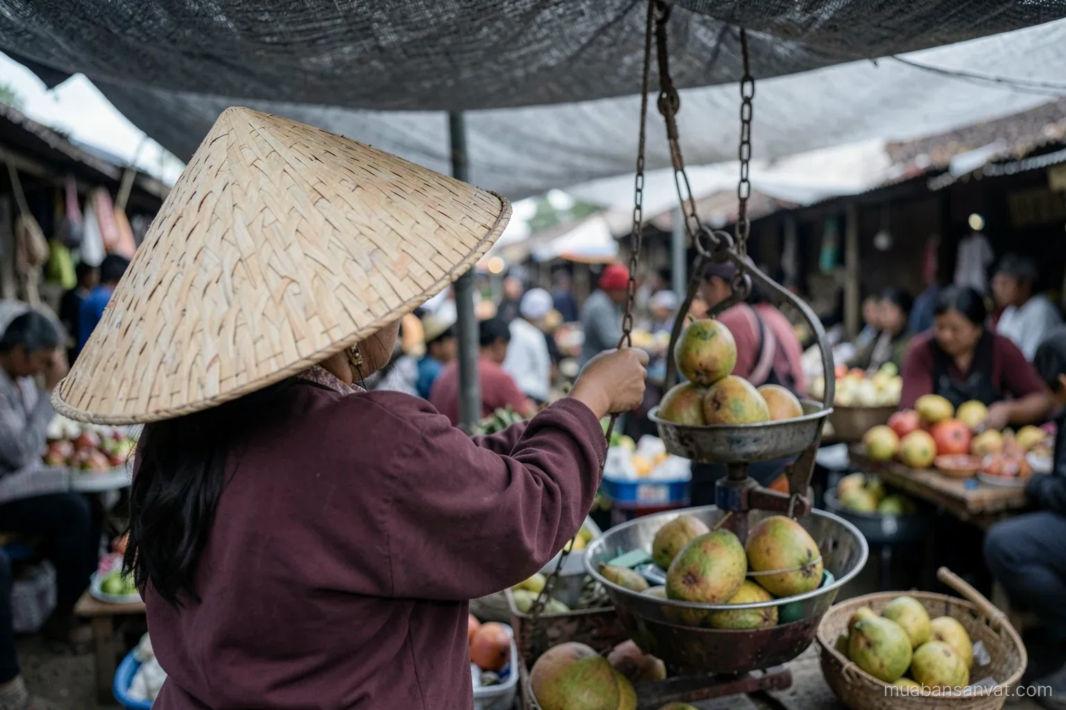 Over-the-shoulder view of a woman in a conical hat weighing pomelos on a hanging hand scale at a small village market under a tarp, diffuse grey light